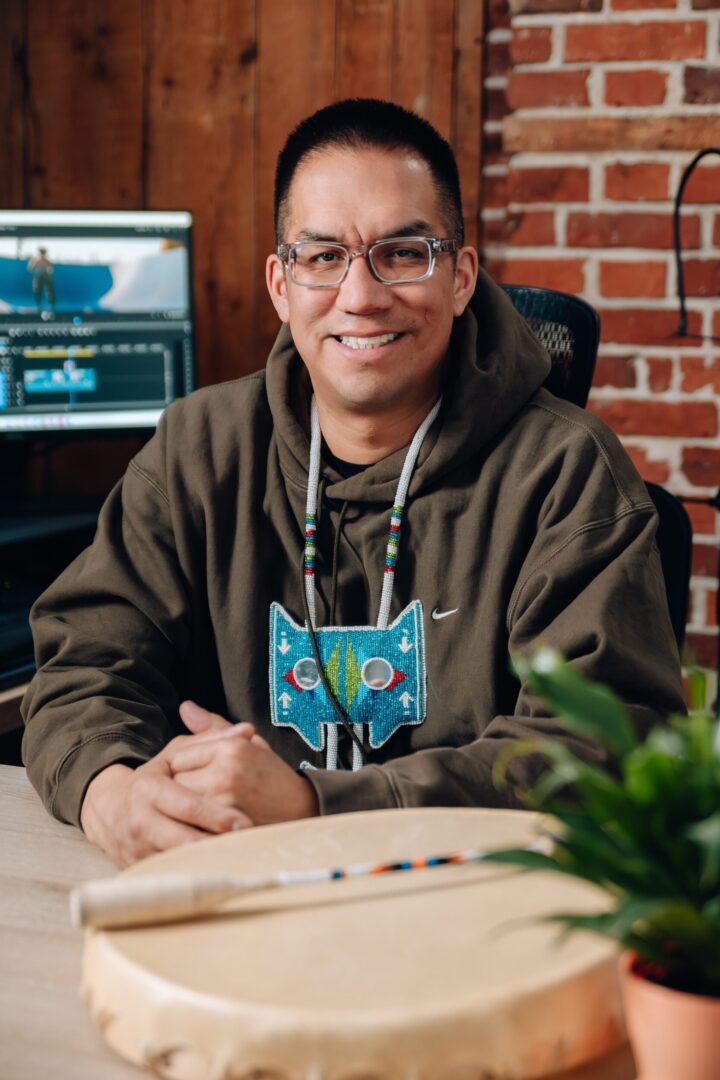 A person sits at a desk, smiling warmly. They're wearing a hoodie with a colorful beaded design. In the foreground is a drum and a plant, while a computer with editing software is visible in the background. The setting is cozy, with wood and brick walls.