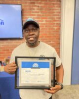 Willis smiles holding his framed certificate from the Maintenance Services Training Program against a brick wall backdrop, conveying pride and achievement.