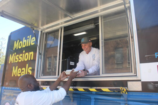 Chef Ric in food trucks smiles and serves a meal to a client. The atmosphere is friendly and supportive.