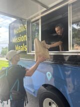 A man in a cap receives a paper bag from a worker inside a blue food truck labeled "Mobile Mission Meals." The mood is positive and giving.