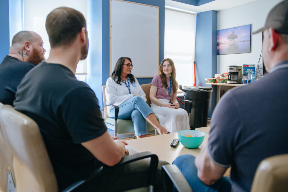 A group of clients sit in a circle in an office setting, engaged in conversation with Mission staff. The room has blue walls, a whiteboard, and a table with snacks.