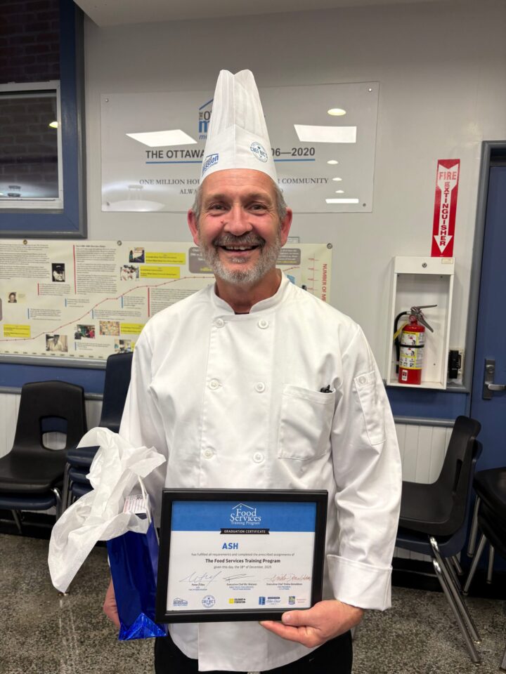 Smiling chef in a white uniform and hat holding a framed certificate and gift bag. The background features a bulletin board and a fire extinguisher.