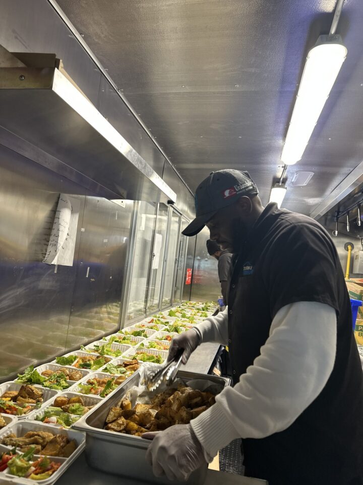 A cook in a black cap and gloves places fried chicken into trays of mixed vegetables in a well-lit kitchen, conveying a sense of focus and efficiency.