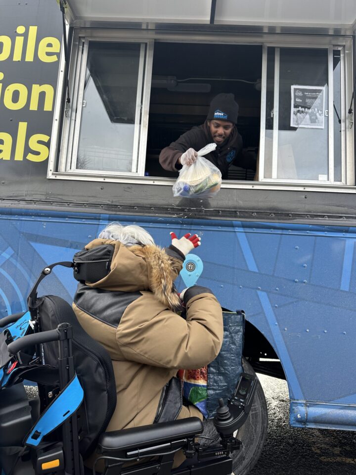 A person in a wheelchair receives a bag of groceries from a food truck worker. The scene conveys care and community support.