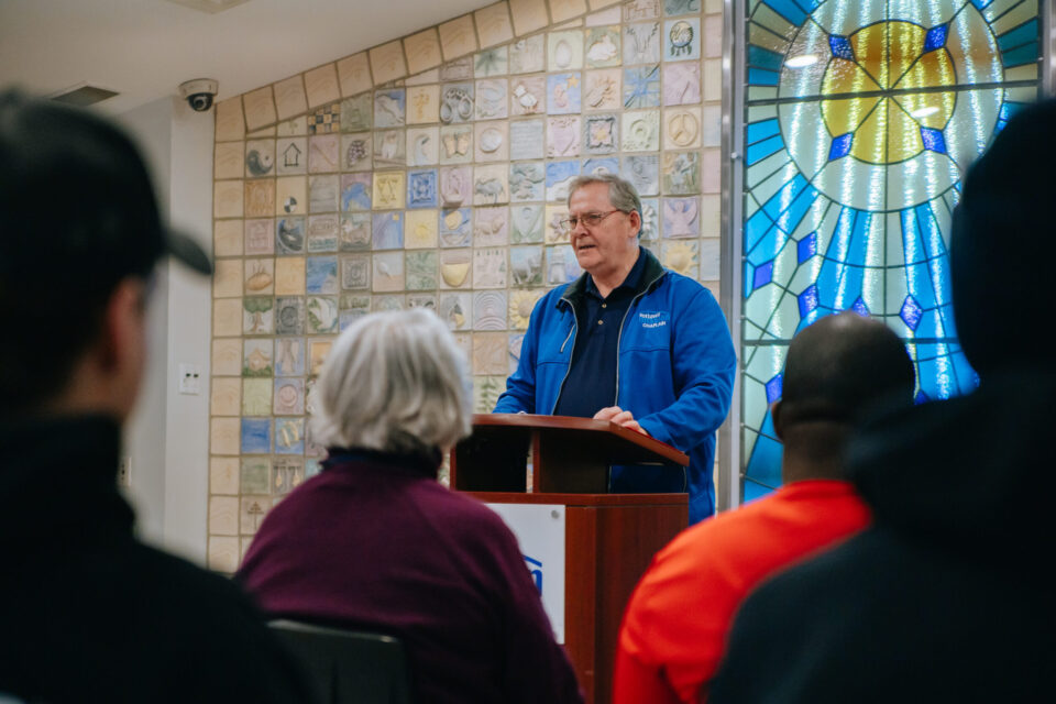 A man in a blue jacket speaks at a podium in front of seated people. Behind him is a colorful stained glass window and a wall with decorative tiles.