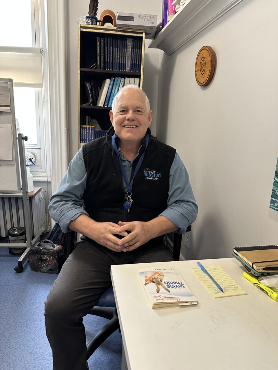 A smiling man wearing a black vest and a lanyard sits at a table with his hands folded in his lap. The atmosphere is warm and inviting.