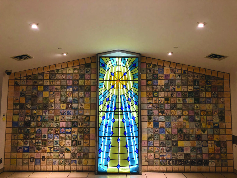 Stained glass window with blue and yellow sun rays at center. Surrounding walls display colorful tile artwork, evoking a sense of warmth and comfort.