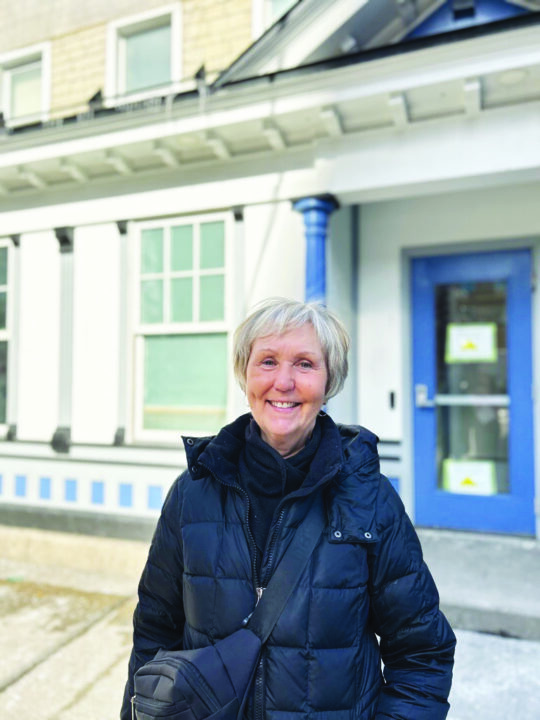 An older woman with short gray hair smiles warmly, wearing a black coat. She stands outside a building with blue accents, white walls, and large windows.