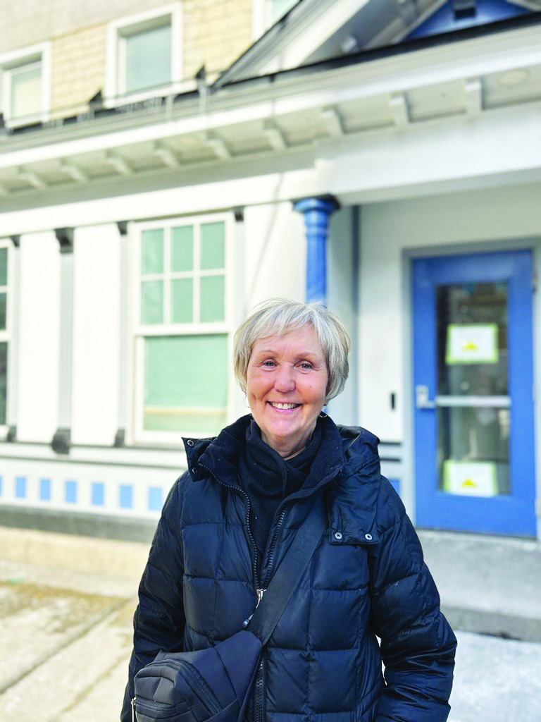 An older woman with short gray hair smiles warmly, wearing a black coat. She stands outside a building with blue accents, white walls, and large windows.