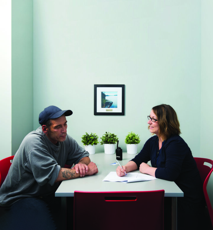 A man looking for help is sitting at the table with a staff member of The Ottawa Mission. She is writing on a sheet of paper and smiling at the man.