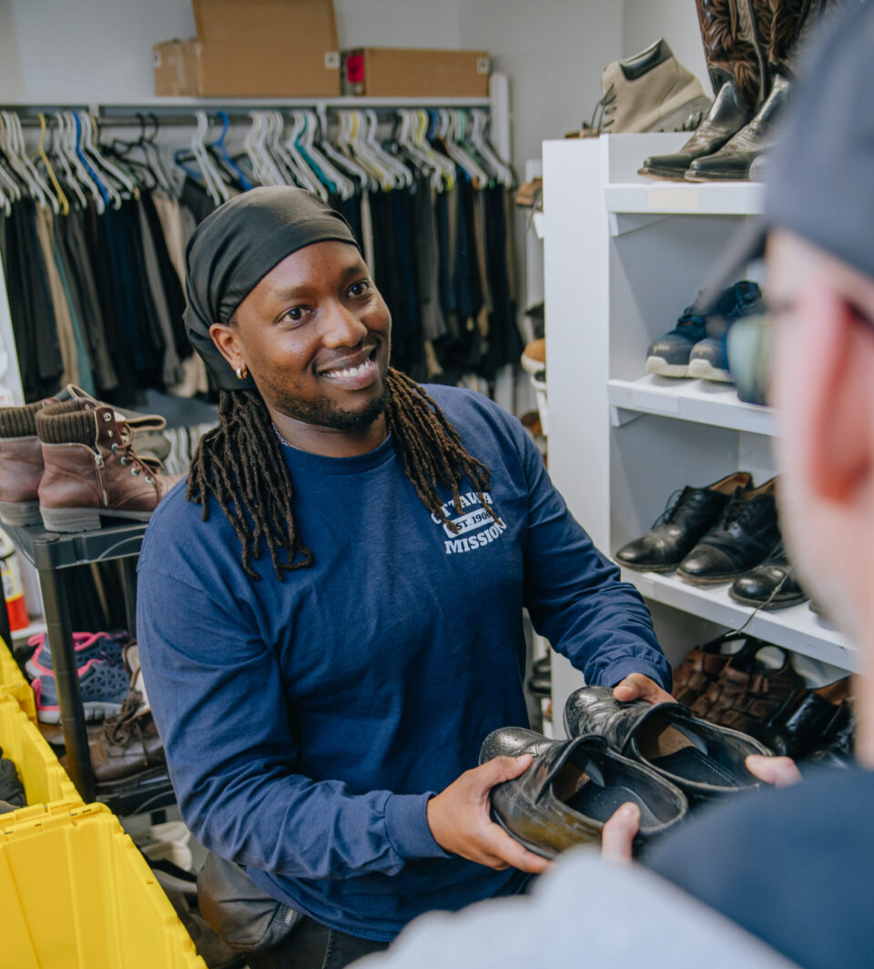 A smiling person in a blue shirt hands a pair of black shoes to a customer in a thrift store. Shelves filled with clothing and shoes are in the background.