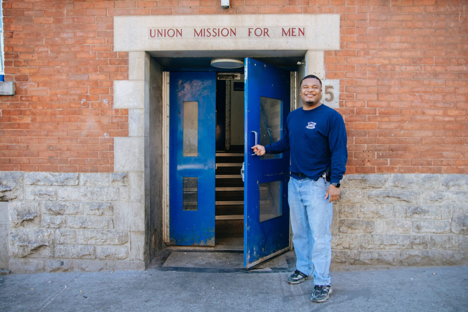 A man in a navy shirt and jeans smiles while holding open a blue door labeled "Union Mission for Men" on a brick building, creating a welcoming tone.