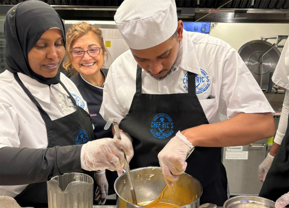 A group of people in a kitchen setting, wearing aprons and gloves, are engaged in cooking. One person is stirring a mixture in a pot while others look on with smiles.