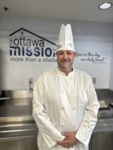 A chef in gala uniform stands in front of a wall with the logo of The Ottawa Mission. He is smiling to the camera.