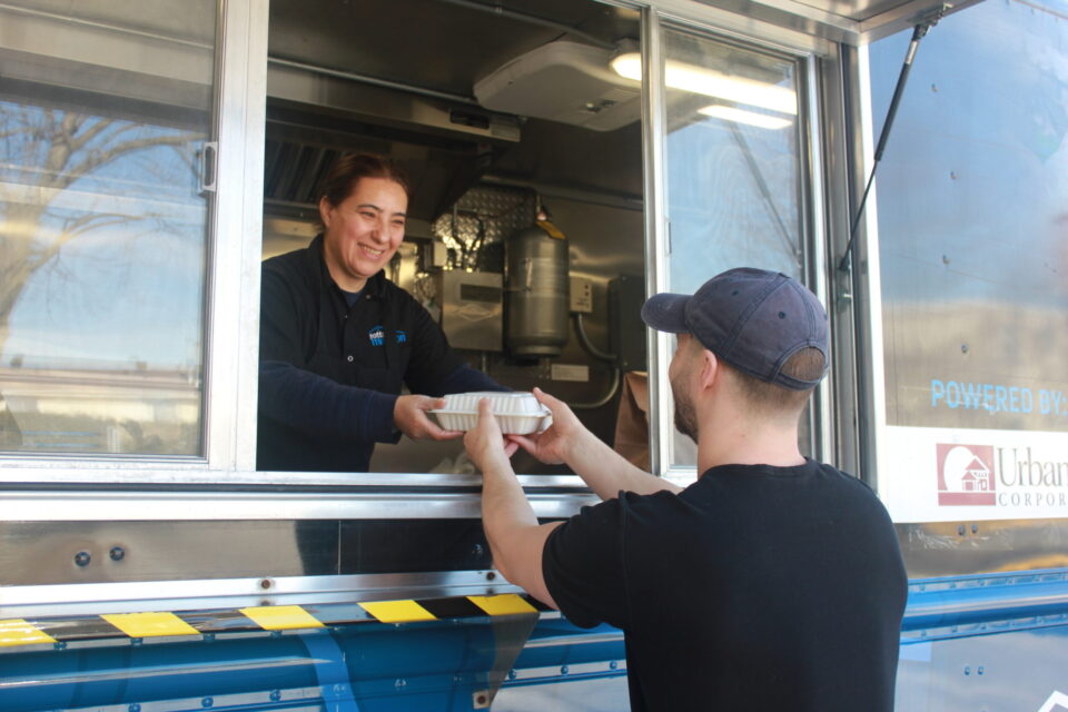 A smiling woman serves food from a "Mobile Mission Meals" truck window to a man wearing a cap. The mood is friendly and supportive.
