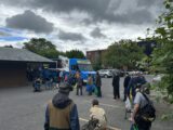 A group of people stand in line at a food truck in a parking lot under a cloudy sky. The scene conveys anticipation and community support.
