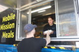 A smiling woman serves food from a "Mobile Mission Meals" truck window to a man wearing a cap. The mood is friendly and supportive.
