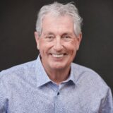 Smiling older man with gray hair wearing a light blue patterned shirt against a dark background, conveying warmth and approachability.