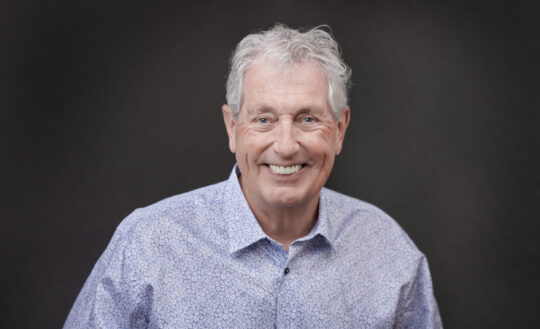 A smiling older man with gray hair wearing a light blue patterned shirt. The background is a soft, dark gradient. The mood is warm and friendly.