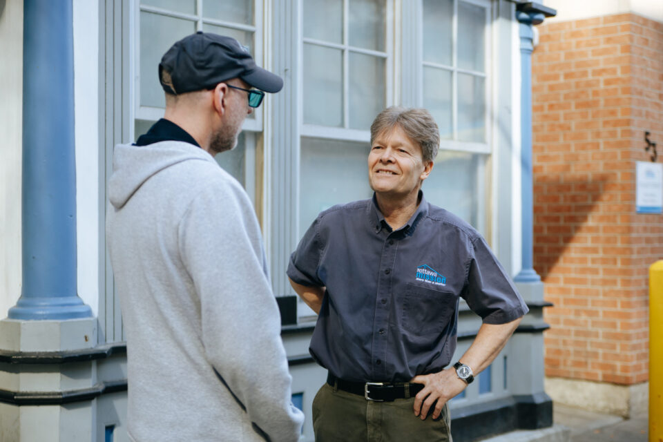 Mission CEO, Peter Tilley has a conversation with a Mission client outside of the building. Peter wears a gray shirt and smiles, while the client, in a cap and hoodie, listens attentively.