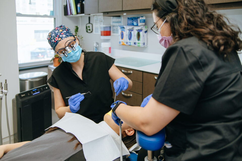 Two dental professionals in masks and gloves perform a procedure on a patient in a modern dental office, conveying a sense of focus and care.