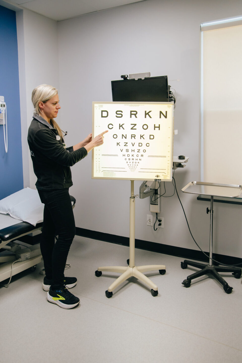 A woman in a medical examination room points to a line on an eye chart. The room is equipped with medical furniture, creating a professional atmosphere.