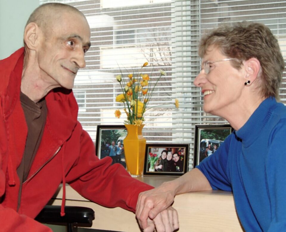 An elderly man in a red jacket and a woman in a blue sweater share a tender moment, smiling at each other and holding hands by a window with blinds. Behind them, a yellow vase with orange flowers and framed photos adorn the table, creating a warm, intimate atmosphere.
