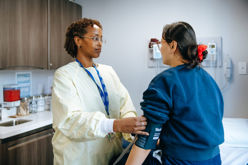 A healthcare professional in a yellow gown checks a patient's blood pressure in a medical office. The environment is calm and attentive.
