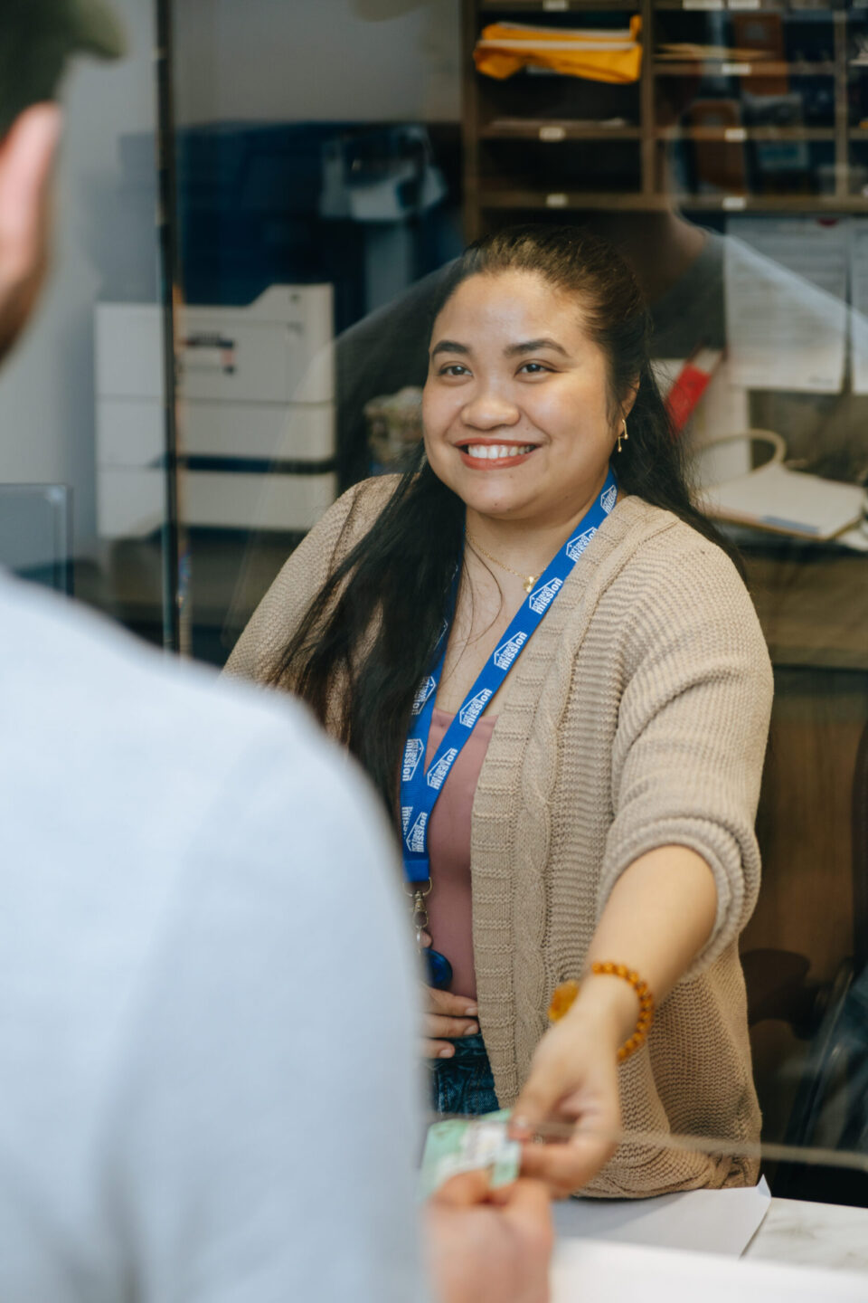 A smiling woman with long dark hair wears a lanyard and cardigan, reaching across a counter to take a health card from an unseen person, conveying friendliness.