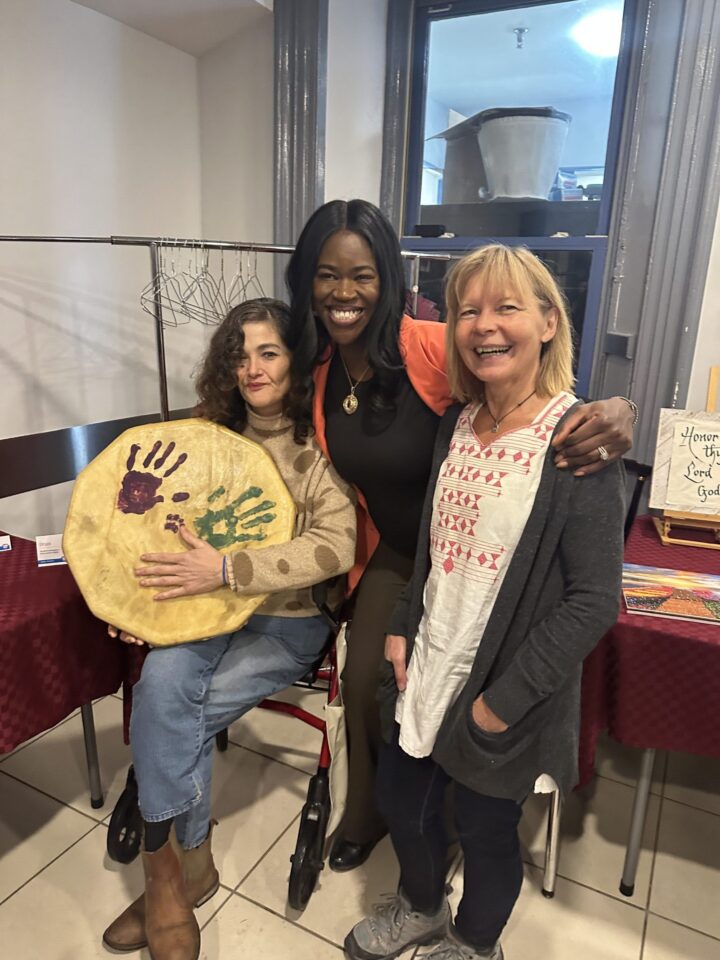 Three women smiling and posing together indoors. One is seated, holding a drum with colorful handprints. The atmosphere is warm and joyful.