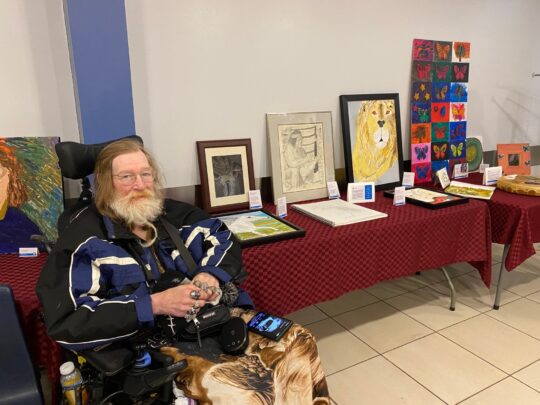 A man with a long beard and glasses sits in a wheelchair, surrounded by various pieces of artwork displayed on tables. He appears to be smiling and is dressed in a blue coat.