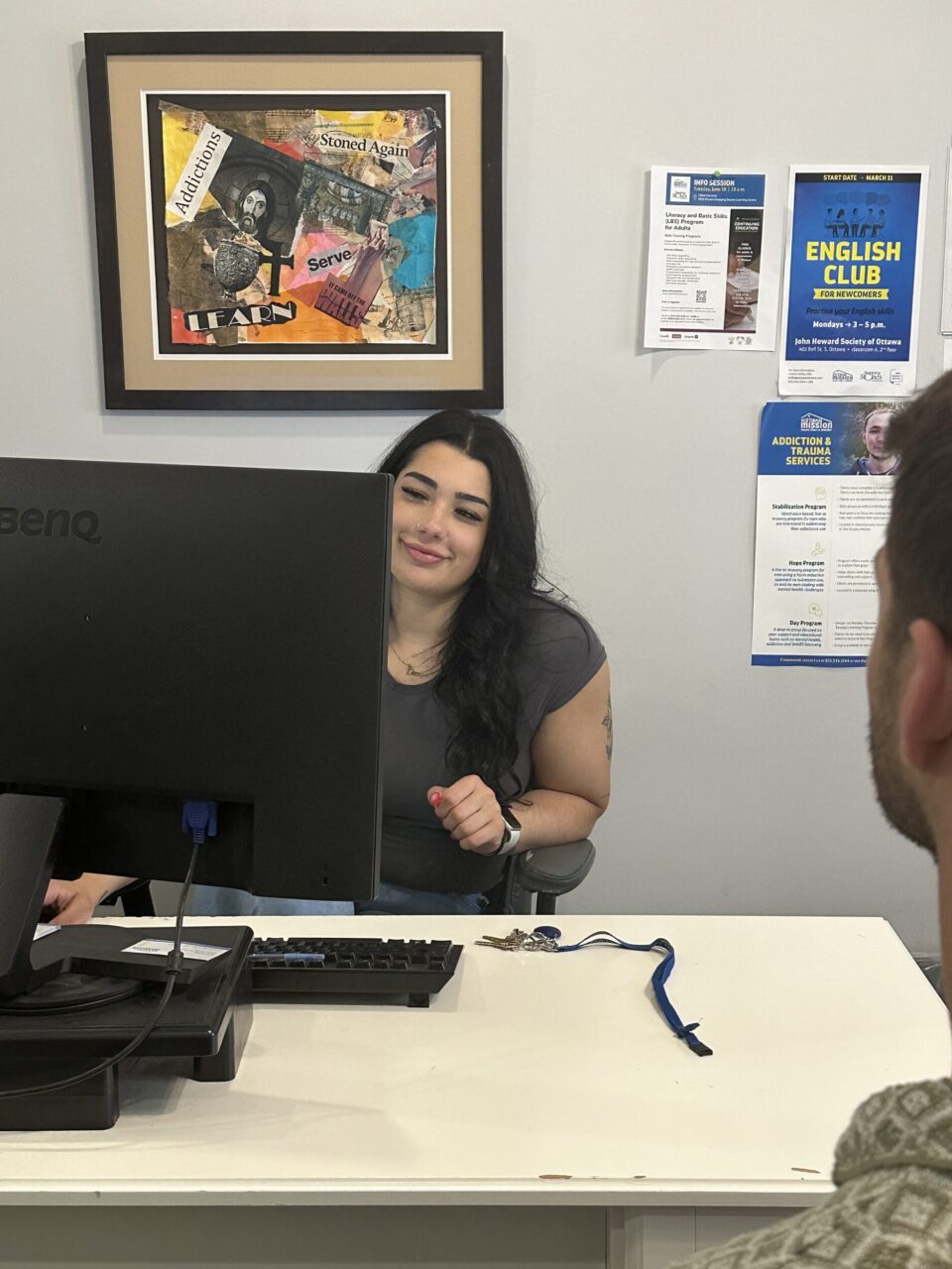 A woman sits at a desk, smiling warmly at a person in a plaid shirt and cap. Papers are spread on the desk, suggesting a friendly conversation.