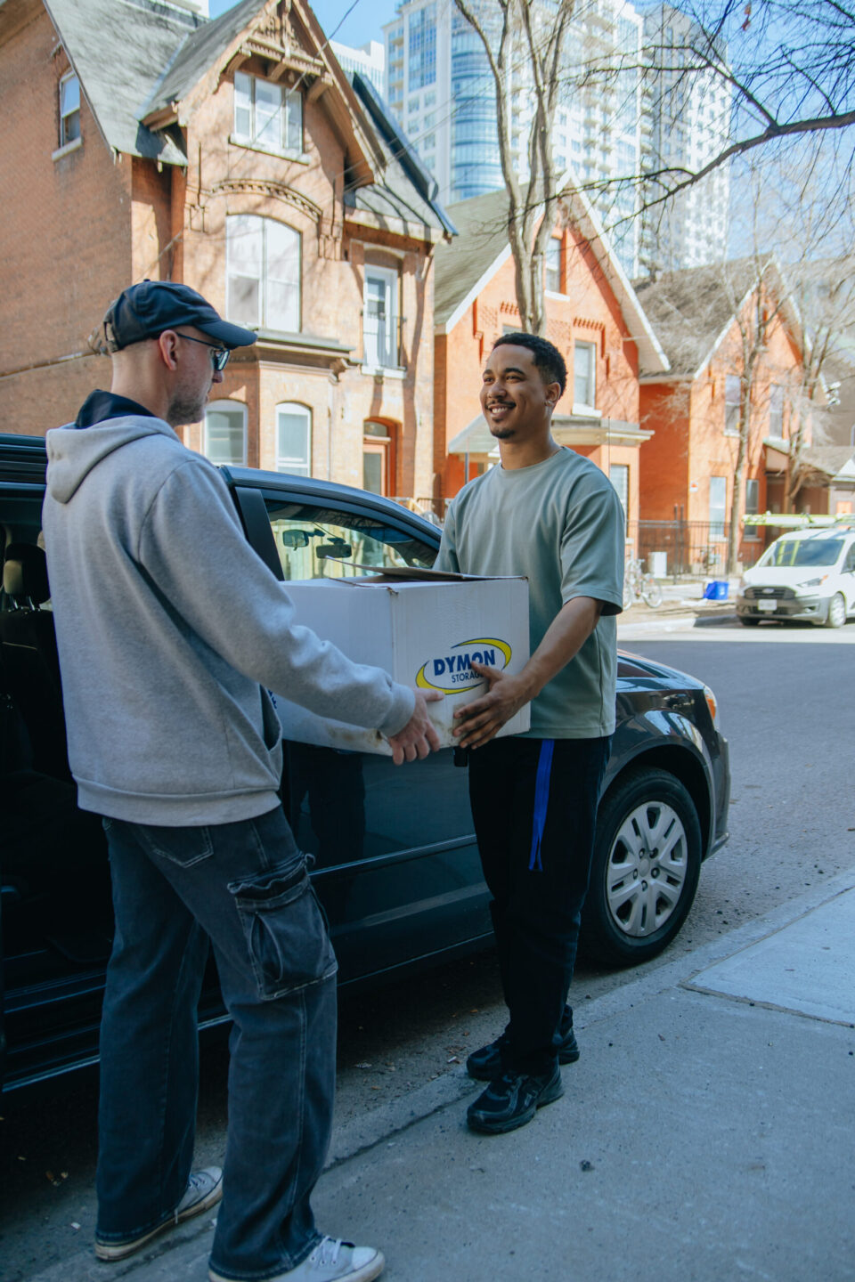 Two men smiling, exchanging a box labeled "Dymon" beside a parked car on a city street. Background shows red brick houses and tall buildings.