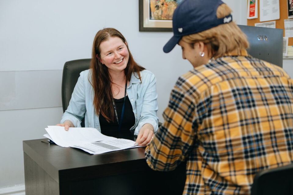 A woman sits at a desk, smiling warmly at a person in a plaid shirt and cap. Papers are spread on the desk, suggesting a friendly conversation.