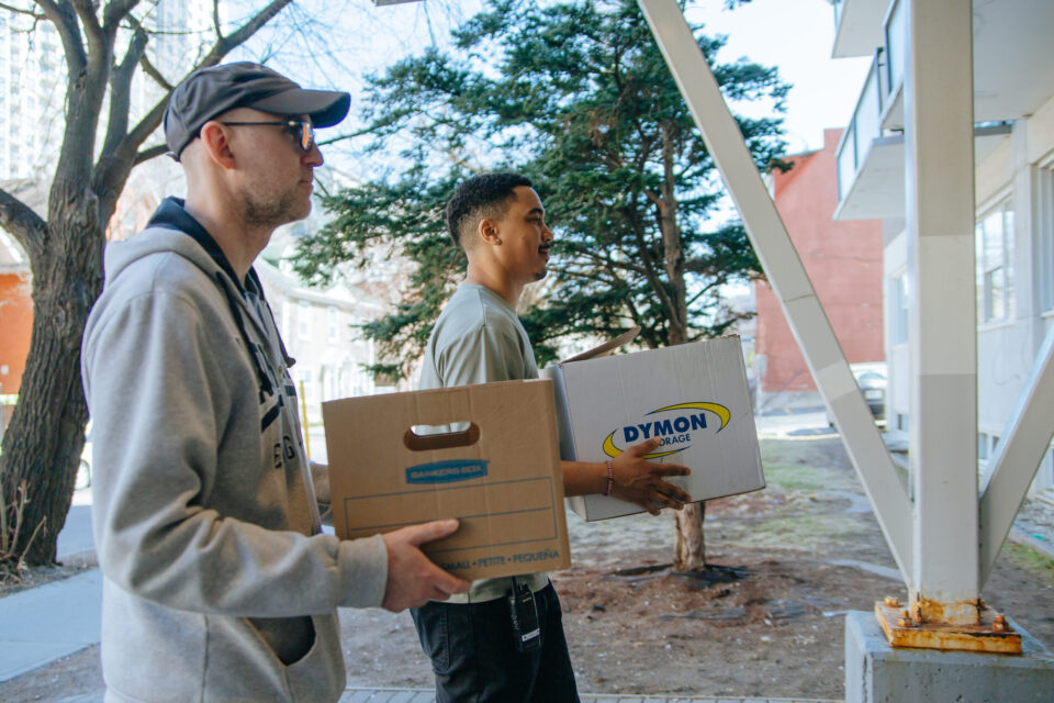 Two people carrying moving boxes approach a building. One holds a "Dymon Storage" box. A tree and urban backdrop create a collaborative, positive mood.