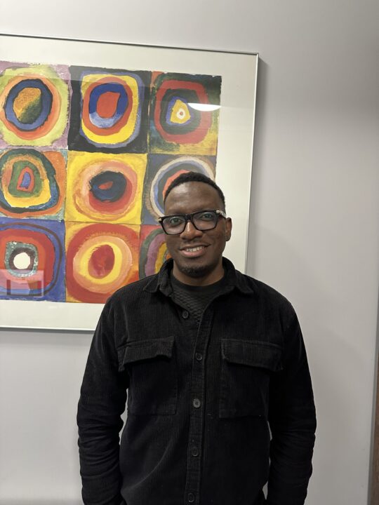 Smiling man in black jacket stands in front of colorful abstract painting featuring concentric circles, creating a vibrant and cheerful scene.
