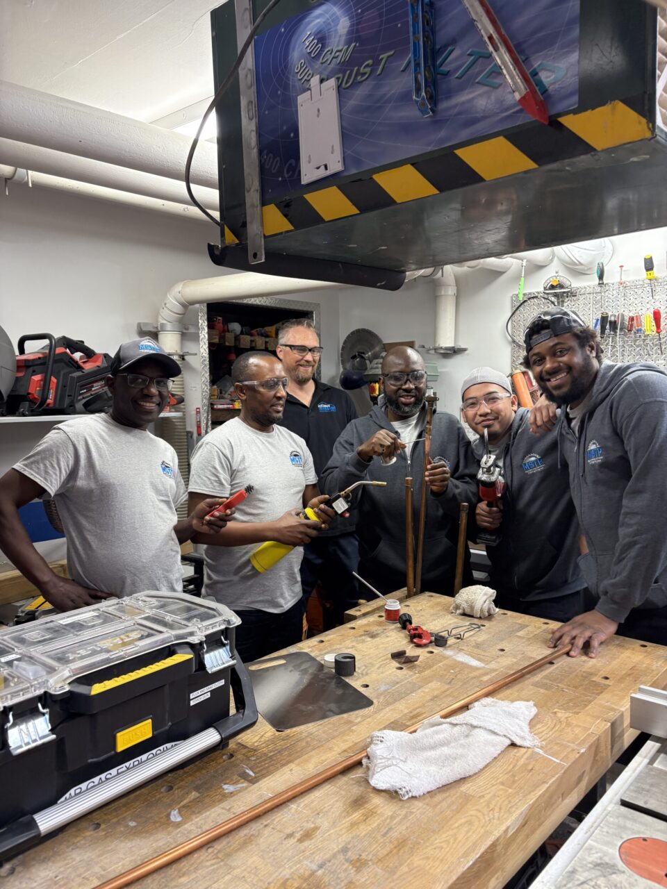Six smiling people stand inside a workshop, holding up copper pipes and tools.