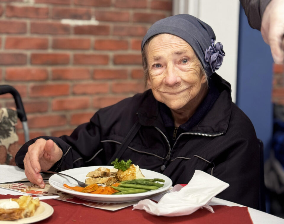 Senior woman wearing a flower hat and a small grin, seated in The Mission dining room ready to eat her meal.