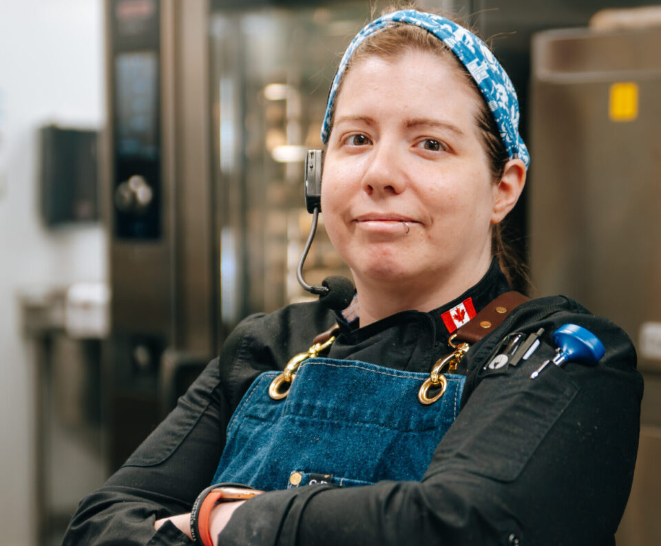 A woman in a blue chef's apron smiles with a determined look on her face inside a commercial kitchen.