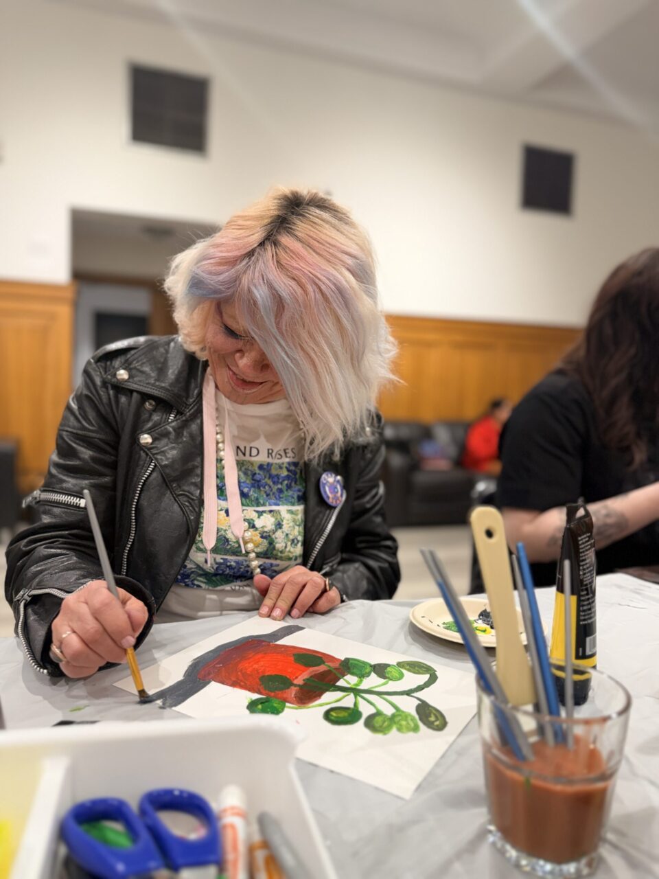 A person with pastel-colored hair wearing a leather jacket is painting on a piece of paper at a table, surrounded by art supplies.