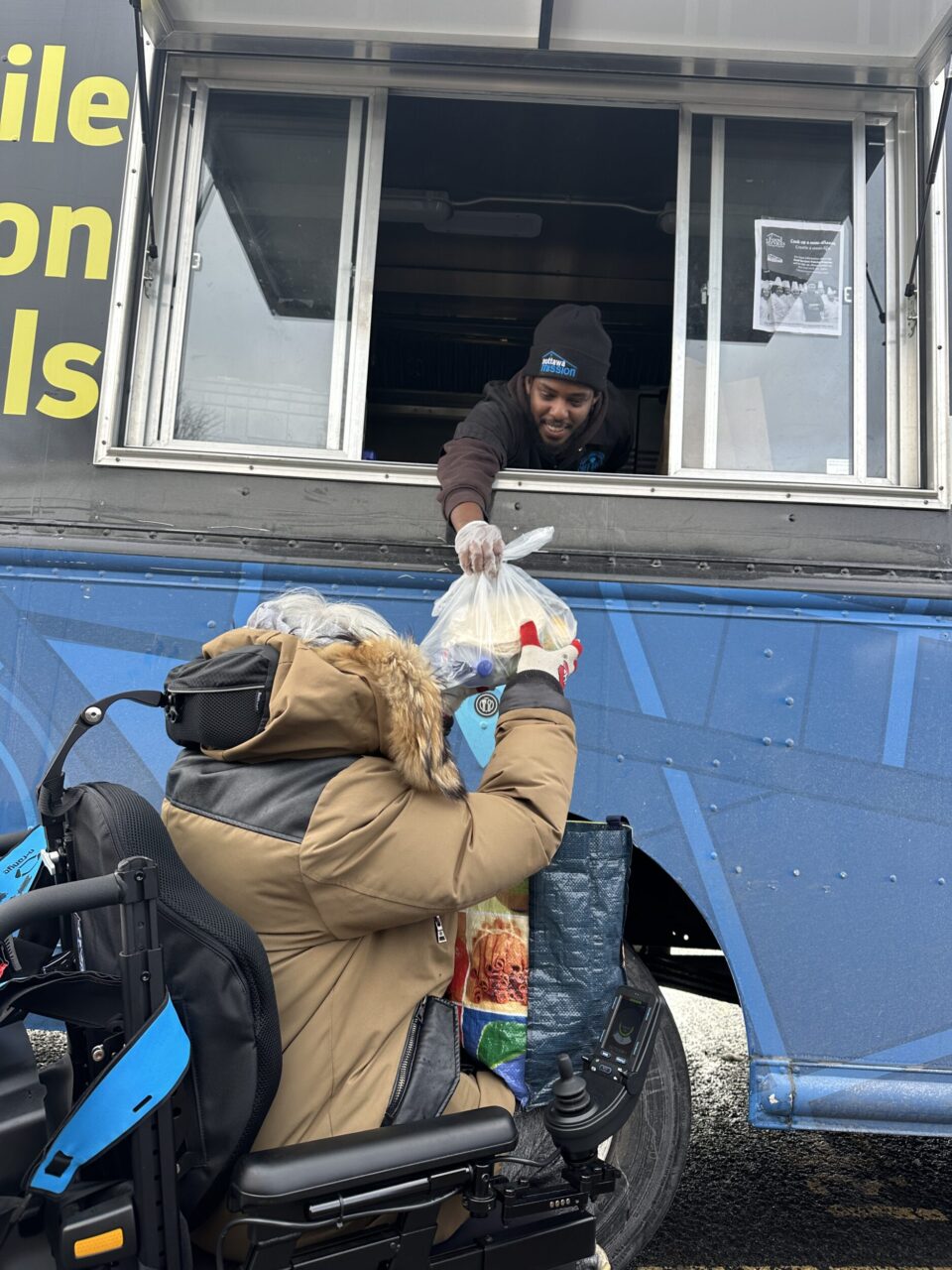 A smiling man hands a to-go meal out of a Mission food truck window to an elderly person in an electronic wheelchair.