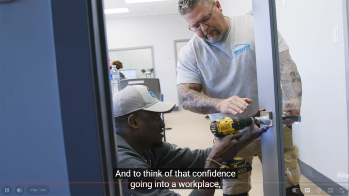 A man with in a grey tshirt helps another man use a power drill on a doorhandle,
