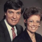 A man in a dark suit and red patterned tie stands beside a woman in glasses and a dark blouse. Both are smiling against a neutral background.