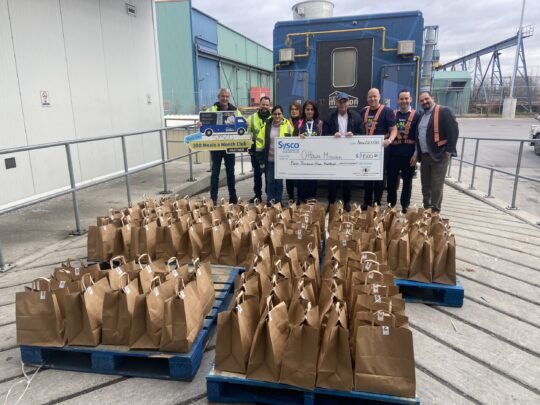 A team of employees stands in front of the food truck with a cheque. In front of them are rows of pre-packaged food truck meals.