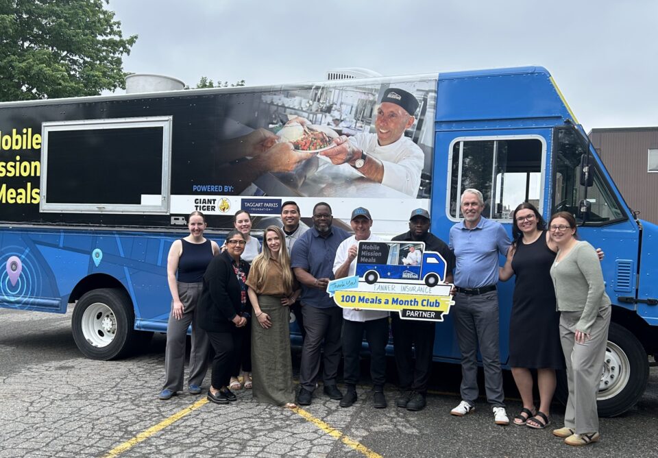 A group of representatives from Tanner Insurance pose with Chef Ric holding a sign of the '100 Meals a Month Club" in front of the food truck.