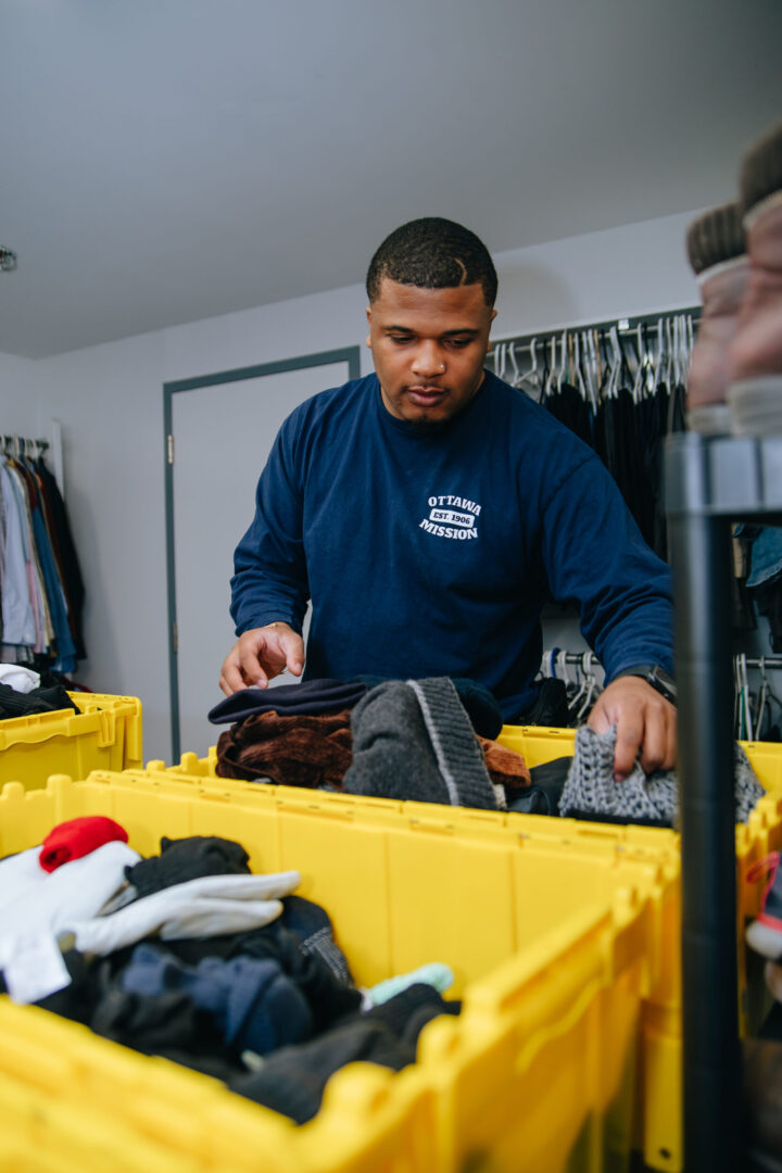 Person in a blue shirt sorting clothes into yellow bins in a room with a clothing rack. The setting suggests a charitable or donation event.