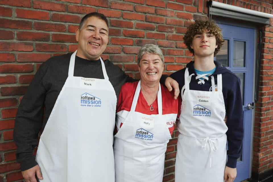Three people wearing aprons stand smiling against a brick wall. The aprons have "Ottawa Mission" printed on them and they are volunteering.