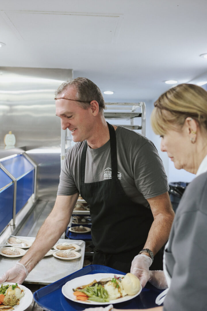 A smiling man in an apron serves a plate of food with vegetables and mashed potatoes in a cafeteria setting, conveying a sense of helpfulness and care.