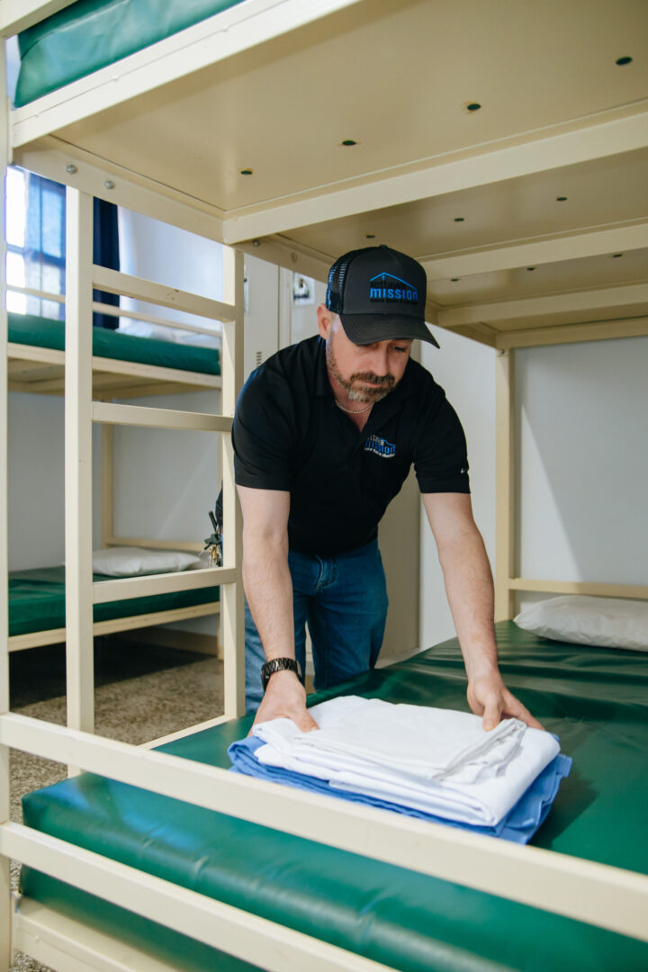 A man in a black shirt and cap arranges folded sheets on a green mattress of a bunk bed in The Mission dorm room, conveying neatness and care.