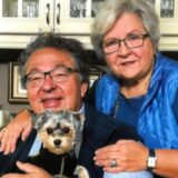 An elderly couple smiles warmly in a cozy kitchen, the man holding a small dog. Both wear glasses and blue clothing, exuding warmth and affection.